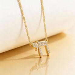 Collier pendentif Chai serti de zirconium en plaqué or et chaîne figaro 2 mm suspendu sur un support laqué blanc beige