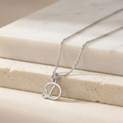 Délicat Collier Initiale Majuscule Cursive en acier inoxydable argent posé sur une surface en marbre beige, hauteur de la lettre 11 mm